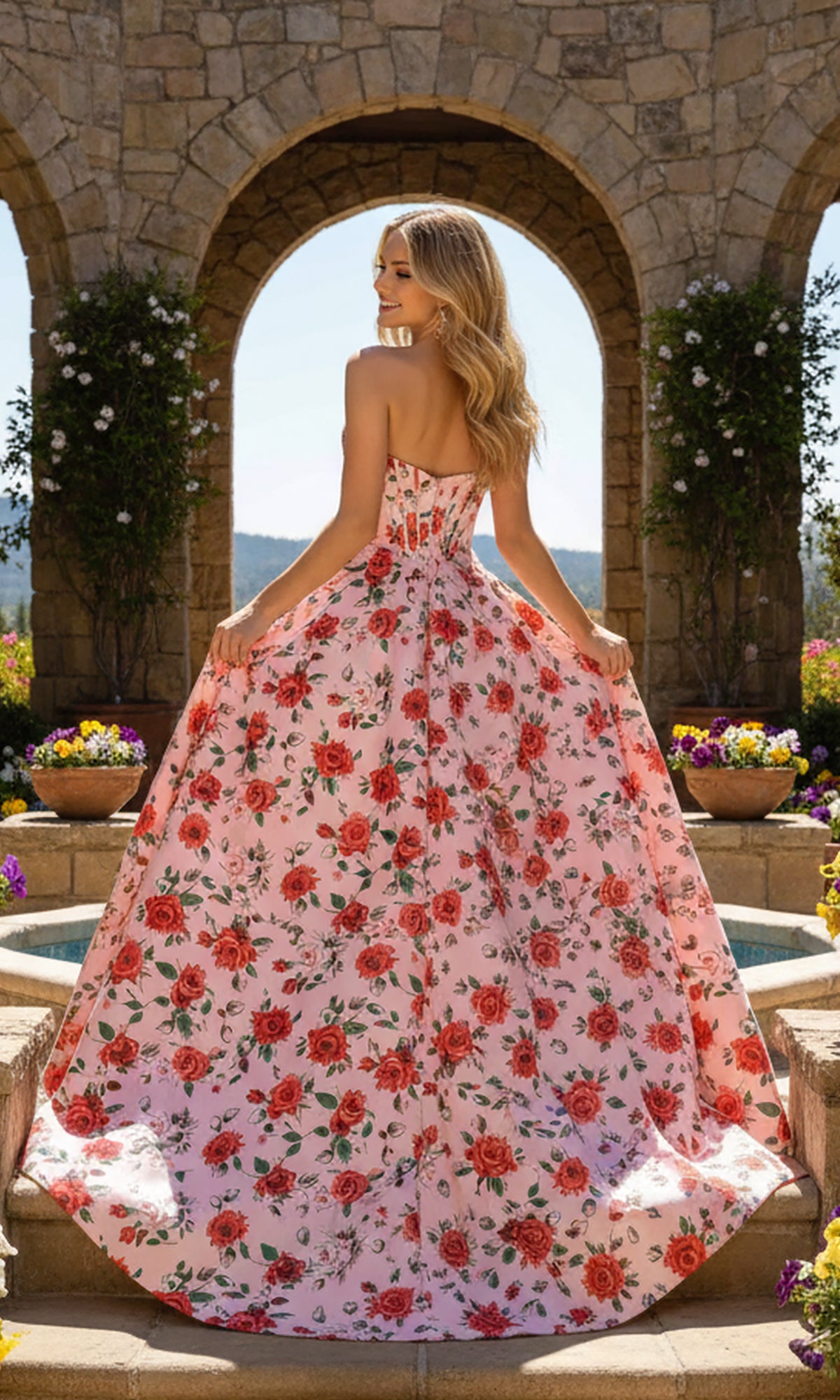 A woman wearing the PromGirl Exclusive 250229 long prom dress poses outdoors before a stone archway, facing away to showcase the strapless pink floral print and corset bodice.